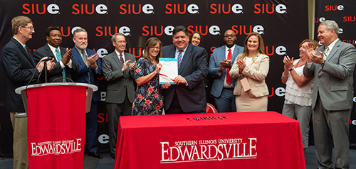 Illinois Governor J.B. Pritzker celebrates the signing of House Bill 2239 during a visit to SIUE’s campus. He stands alongside Rep. Katie Stuart and is surrounded by members of the SIU Board of Trustees and additional contributing legislators.