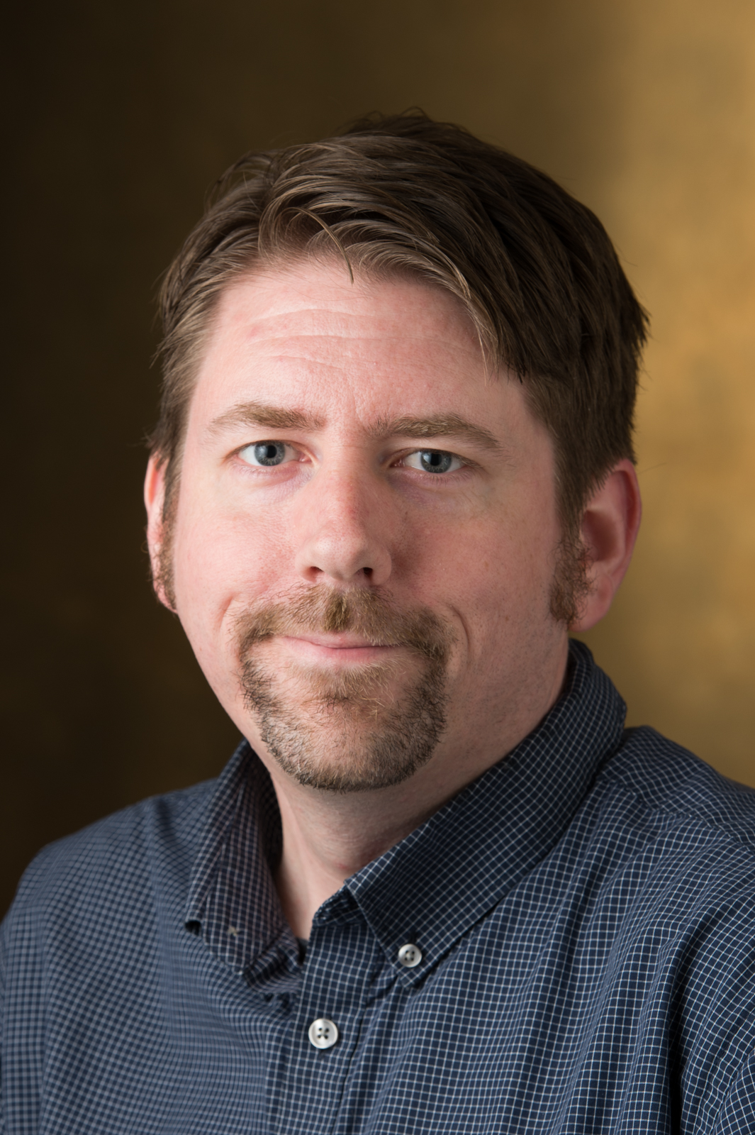 A portrait of Andy Koch, a middle-aged, fair-complected man. Andy is smiling and wearing a plaid shirt.