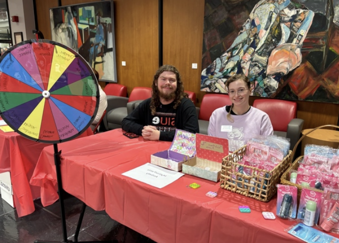 1 EOA practicum student and 1 volunteer sitting behind the EOA table with baskets of prizes and a prize wheel 