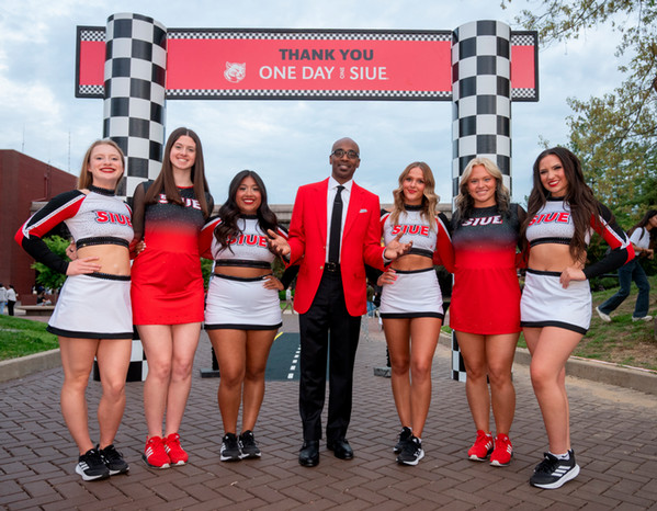Group of SIUE cheerleaders and Chancellor Minor in a red blazer stand beneath a Thank You One Day One SIUE checkered banner on campus.