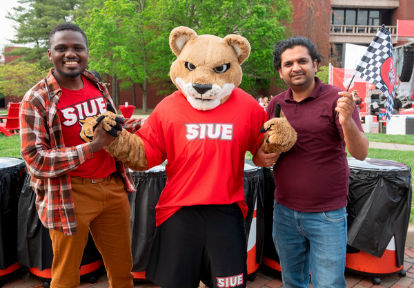 Two attendees pose with the SIUE cougar mascot during the One Day One SIUE event, one holding a checkered flag.