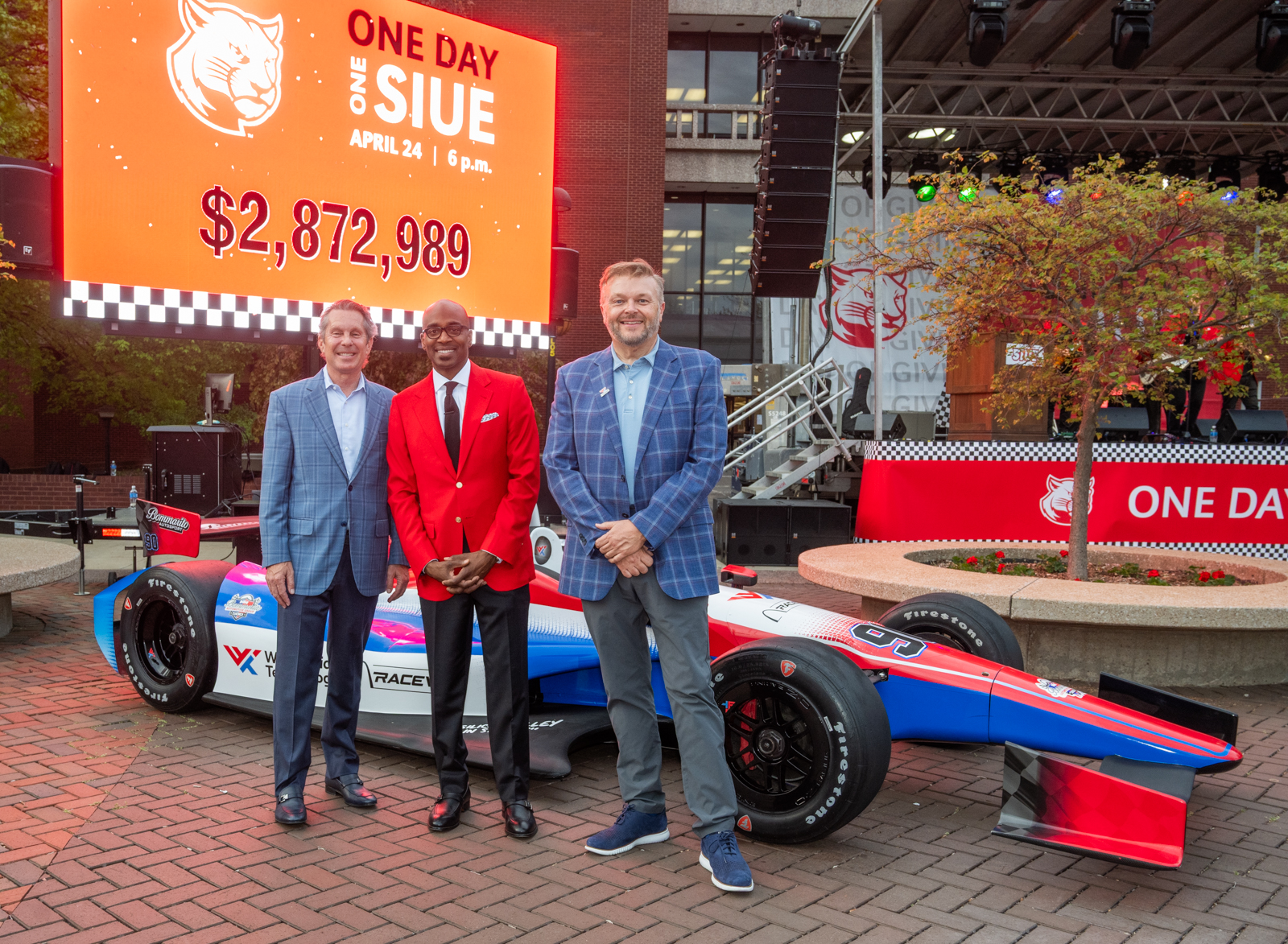 Three men pose in front of a race car display at One Day One SIUE with a large fundraising total shown on a screen behind them.