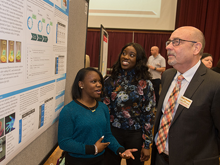 URCA Assistants Daria Whitted (front) and Darzanae Crite describe their research to College of Arts and Sciences' Dean Greg Budzban.