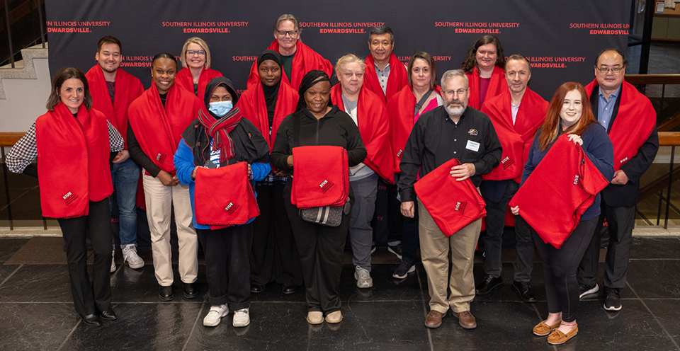 Sixteen faculty and staff members of SIUE pose with red fleece blankets in the Goshen Lounge