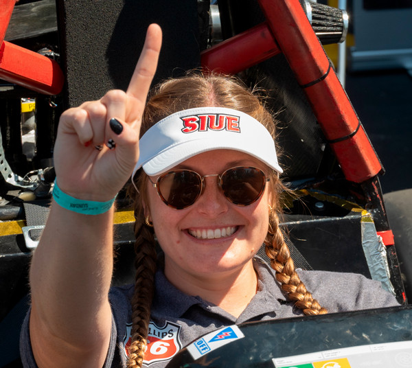 Student engineer with braided hair and sunglasses, sitting in the drivers seat of a Cougar-built racecar holding her hand in a number one gesture at World Wide Technology Raceway on race day
