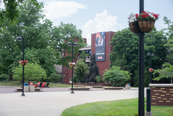 Landscape photo of the Stratton Quadrangle on a Summer cloudy day featuring hanging baskets affixed to the light posts with red and white geraniums. The view is looking toward the Lovejoy Library from Builders of University Plaza
