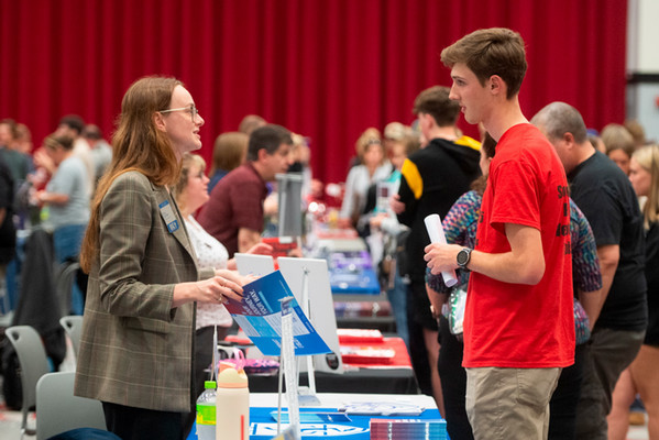 Photo of a prospective student interacting with a person behind a table at a college fair and handing that young student a folder of information