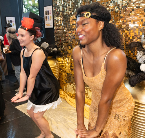 Photo of SIUE Theater and Dance Students dancing in front of a gold shimmering display wearing flapper dresses and vintage mary jane dance shoes. They are performing a variation of the Charleston