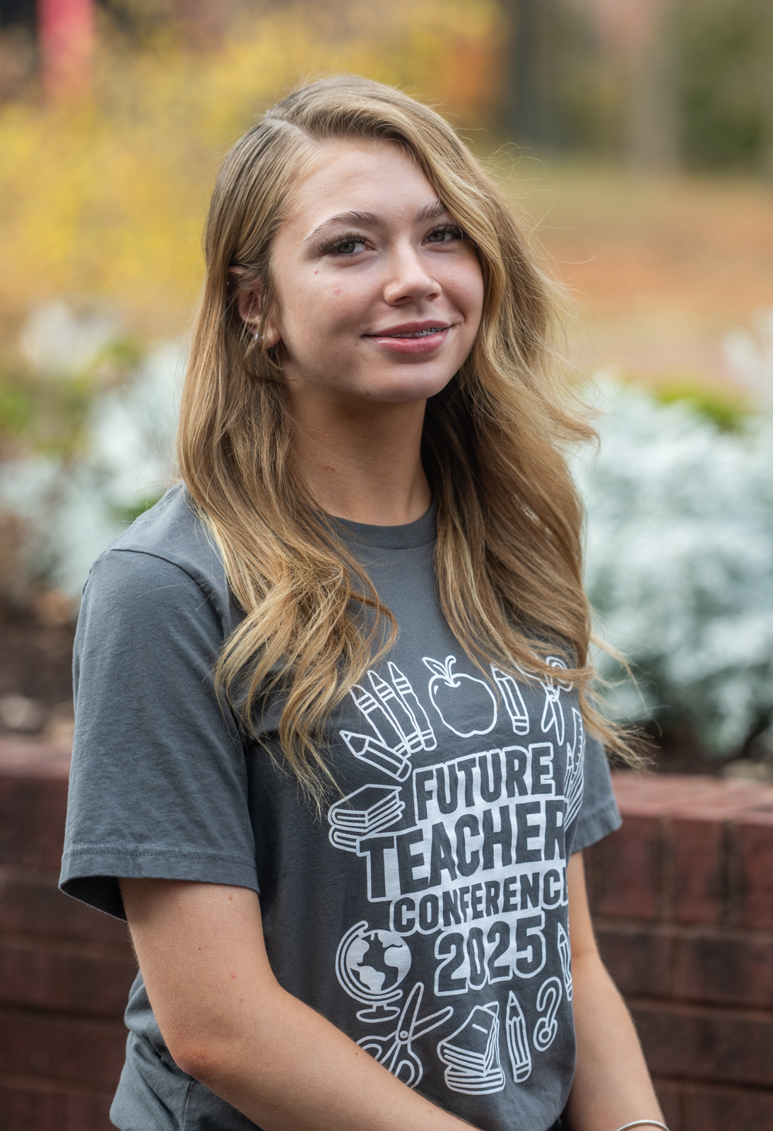 Portrait of a student wearing a gray ‘Future Teacher Conference 2025’ T-shirt, standing outdoors with soft autumn colors in the background.