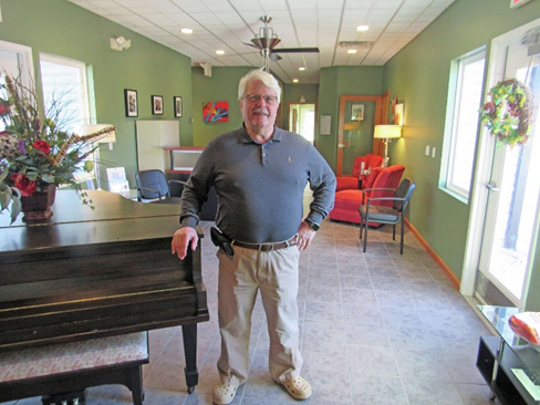 Photo of Larry Lexow in a living room waiting area standing next to a piano an outside door decorated with a holiday wreath