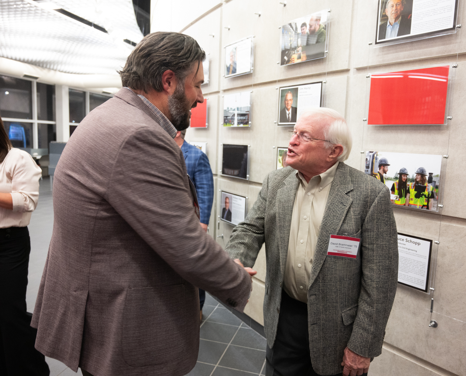 Photo of Jonathan Fowler shaking hands with David Brammemeir in front of the Wall of Fame in the Atrium of the Engineering Building