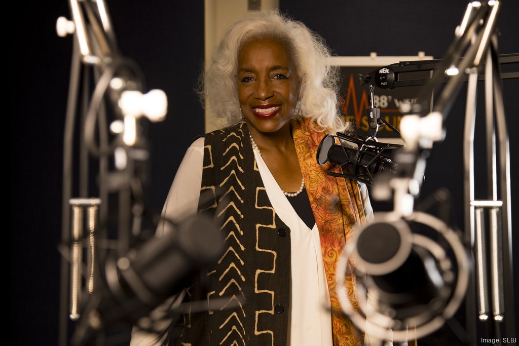 A woman stands smiling in a radio studio, surrounded by microphones and recording equipment.