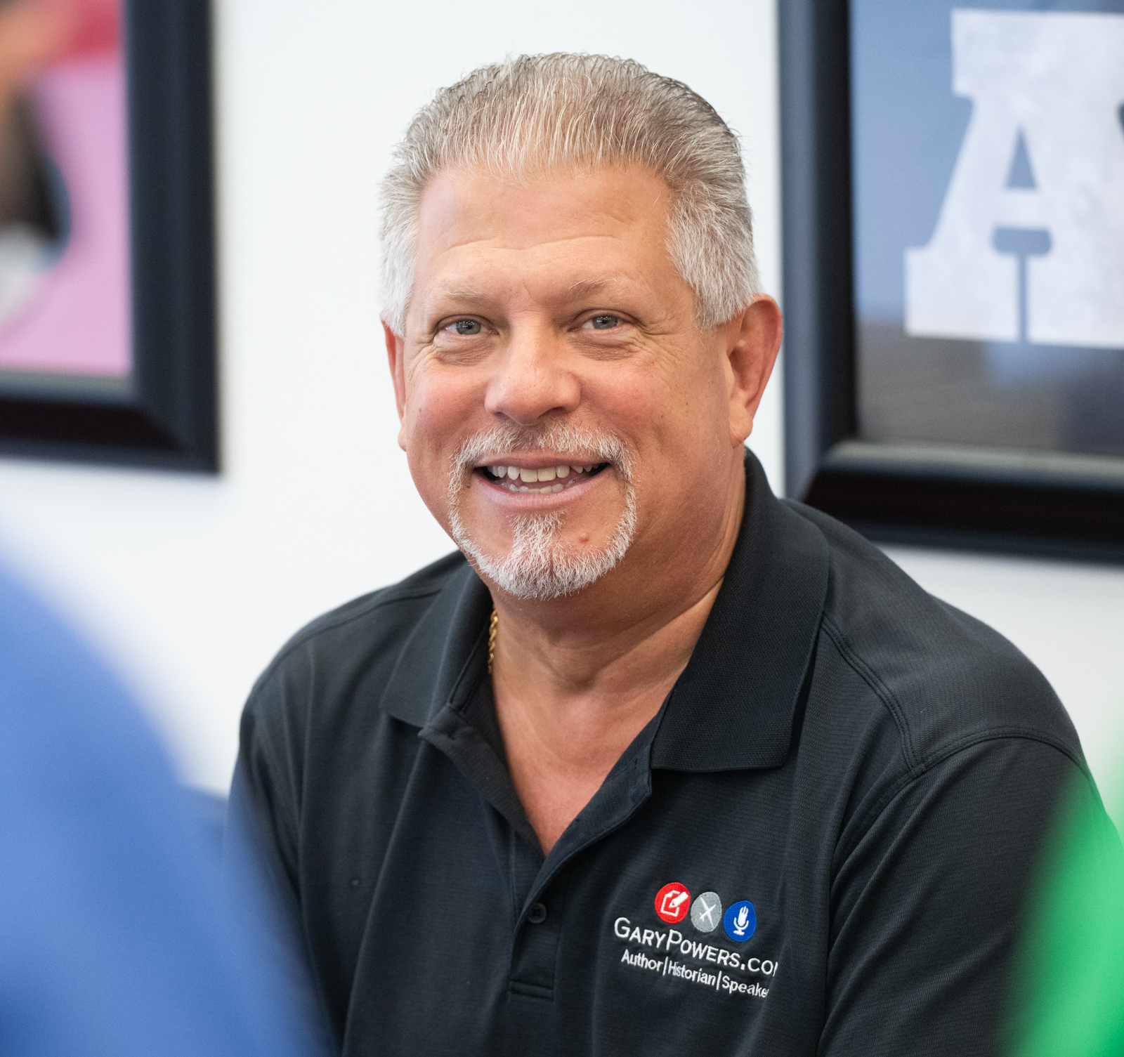 “A smiling man with short gray hair and a goatee sits indoors wearing a black polo shirt with author, speaker, and historian logo for Francis Gary Powers. Framed artwork hangs on the wall behind him.”