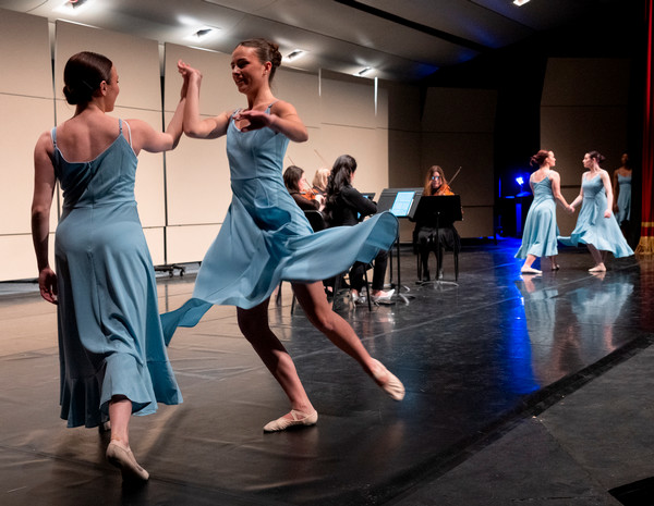 Dancers in blue costume dresses dance in pairs on stage with orchestra