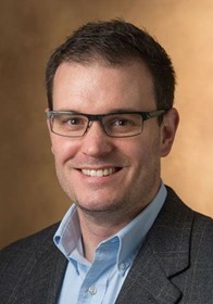 Jeffrey Manuel poses for a professional headshot wearing glasses, a collared shirt, and a blazer against a neutral background.
