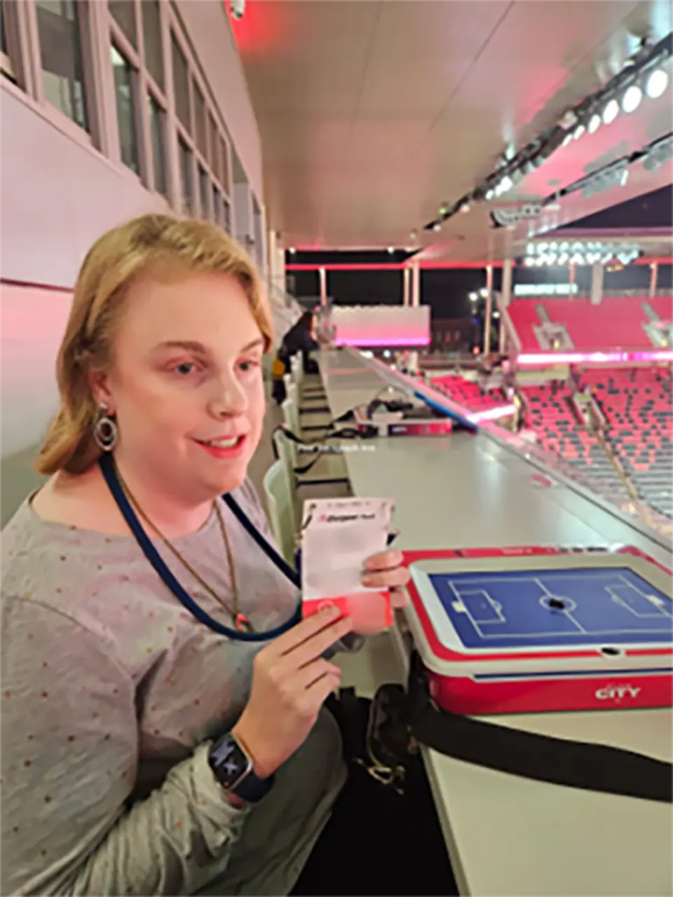 Lacey Weissenborn sits in the press area at CITYPARK holding a media credential, with a tactile play board in front of her used to follow and report on the game.