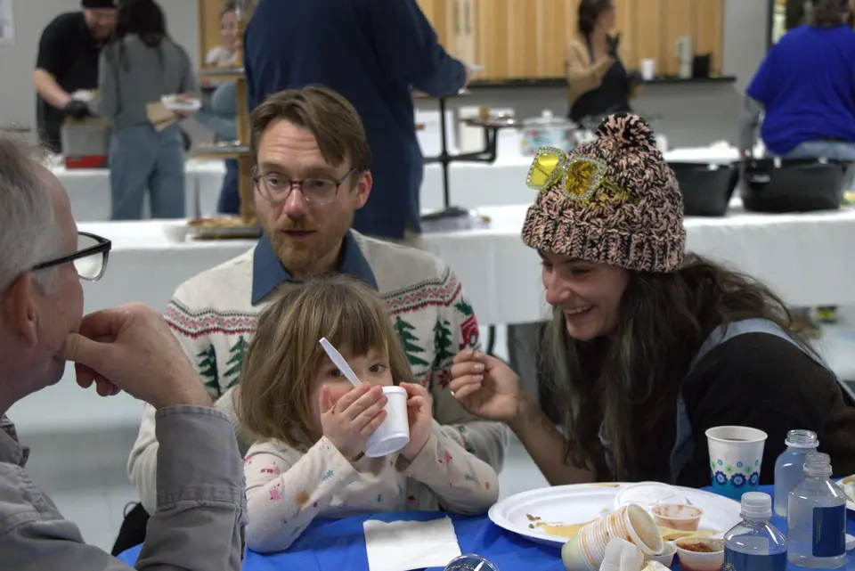 Community members, including a child eating soup, sit together at a table during an Empty Bowls meal event, with serving tables and volunteers visible in the background.