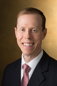 Professional headshot of Paul Rose smiling in a suit and tie against a warm brown studio background.