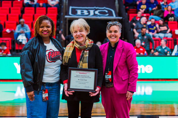 Three women face camera on basketball court while one holds award