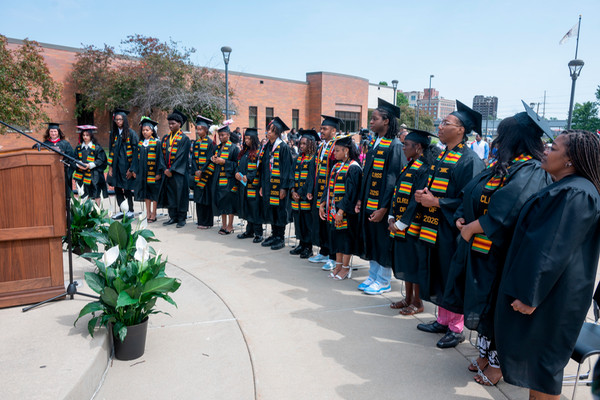 Graduates wearing black caps and gowns with colorful stoles stand together during the SIUE East St. Louis Charter High School Commencement ceremony outdoors on a sunny day. A speaker stands at a podium as the Class of 2026 listens  and stands