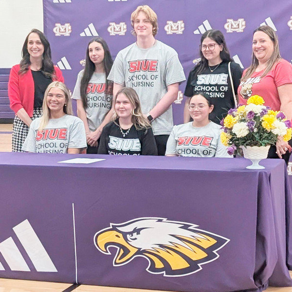 Students wearing SIUE School of Nursing shirts pose during a signing event at Civic Memorial High School in Bethalto alongside Jessica Kerr, far left, and Angela Andrews, far right. The group stands behind a table with the Civic Memorial Eagles logo 