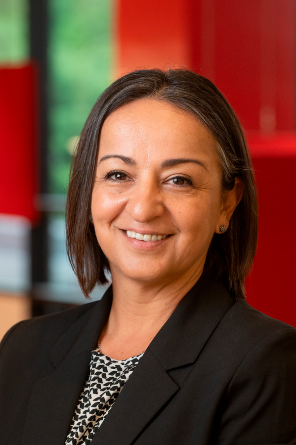 Portrait of Dzenana Mruckovski smiling and wearing a black blazer over a patterned top against a softly blurred red and green background.