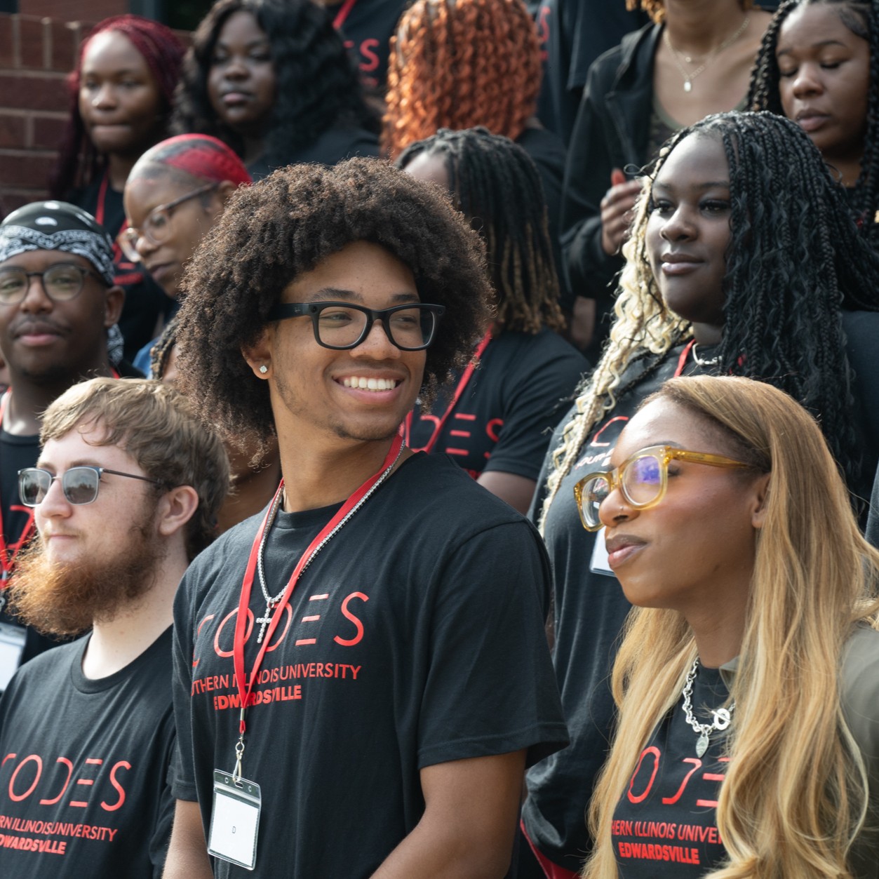 A young man stands amongst a group looking reflective and enjoying the afternoon. Everyone in the group is wearing matching Codes t shirts