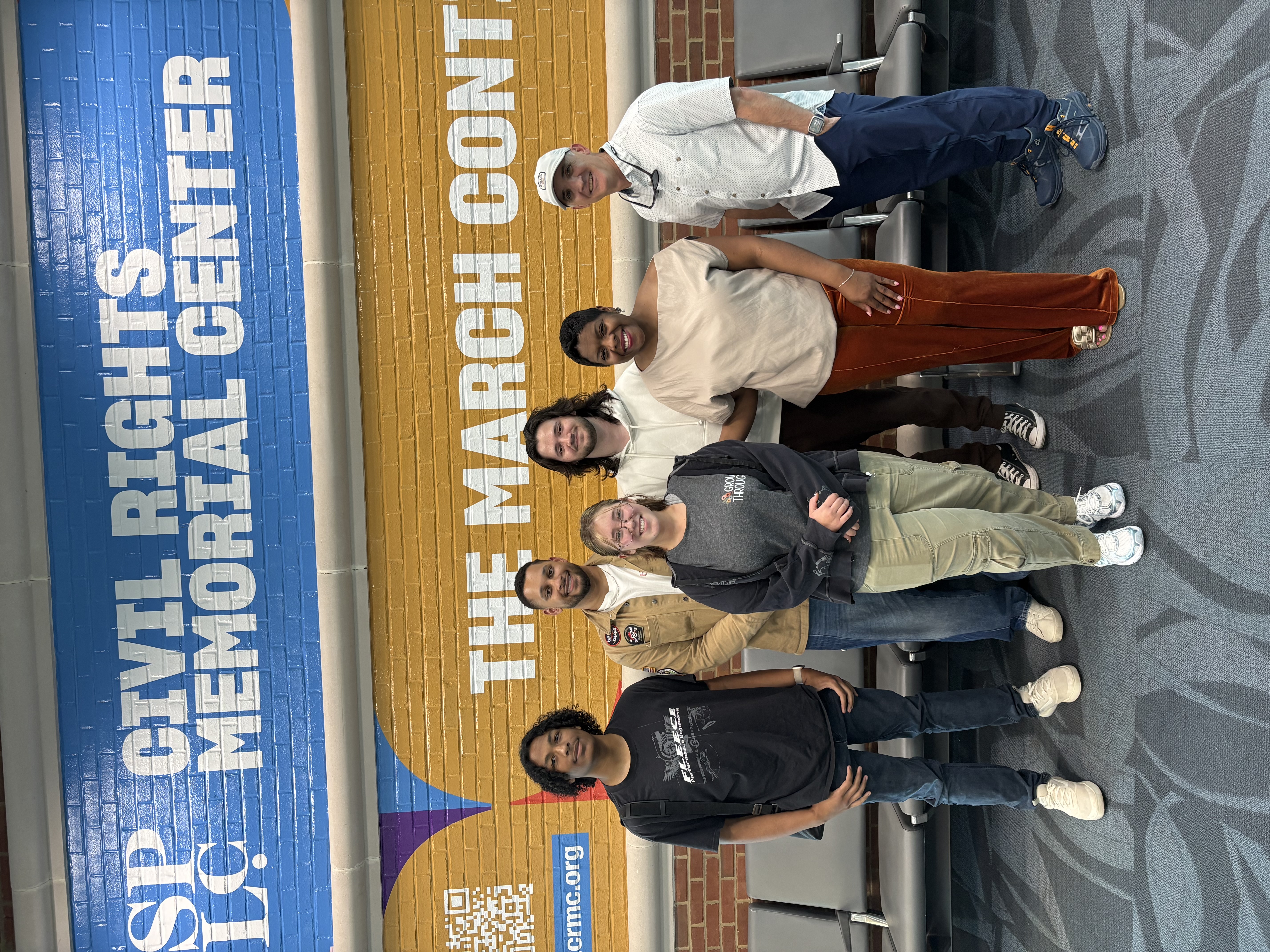 Group shot of six people, including Tandra Taylor, Brian Jack, and four students standing in front of a row of seats at the Civil Rights Memorial Center in Montgomery. The brick wall behind them is pained brightly with the words "the March Continues"