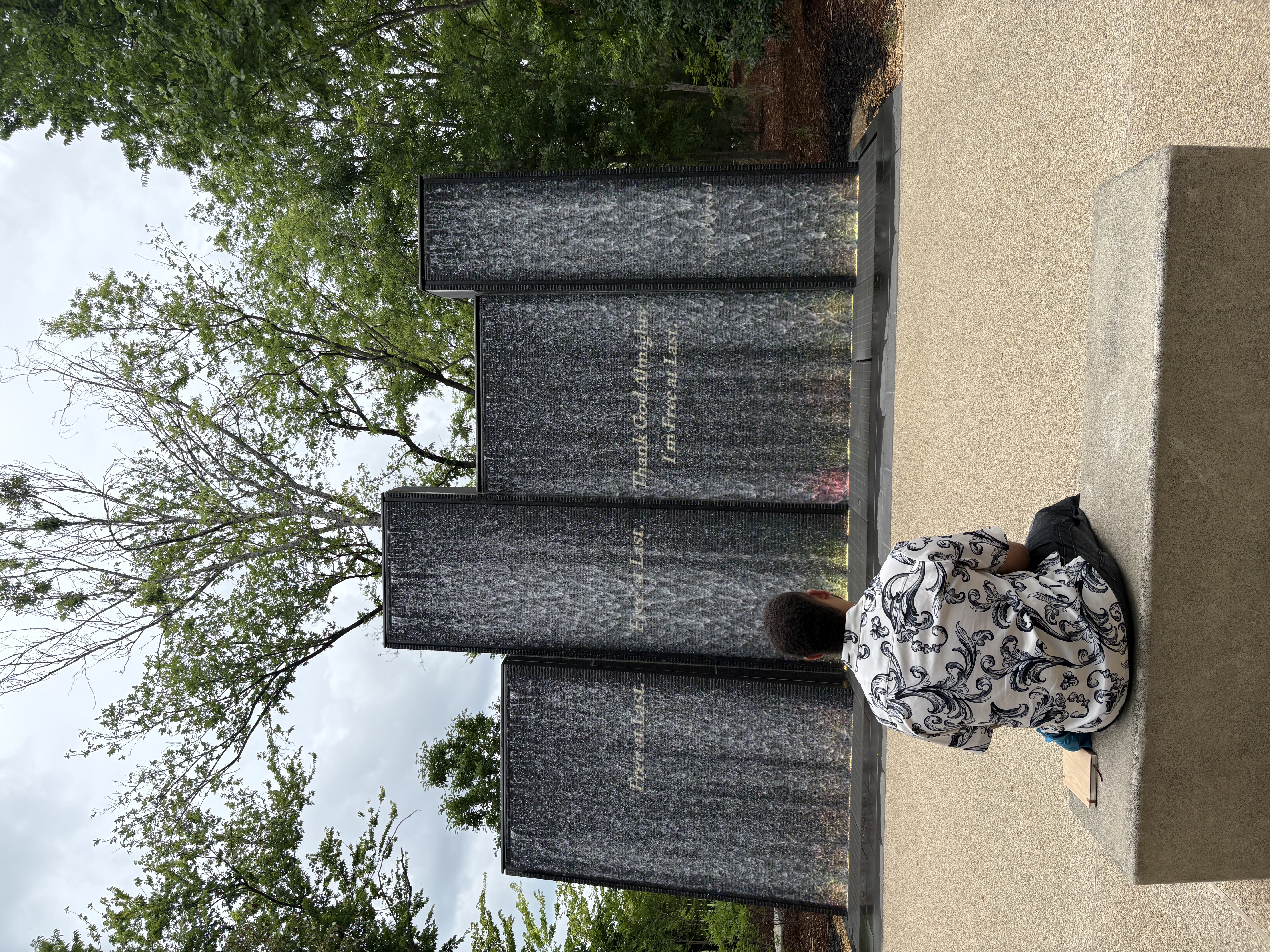 Photo of a student taken from behind. He sits on a marble bench in a sculpture park pondering four large rectangular monuments that resemble buildings in front of him. 