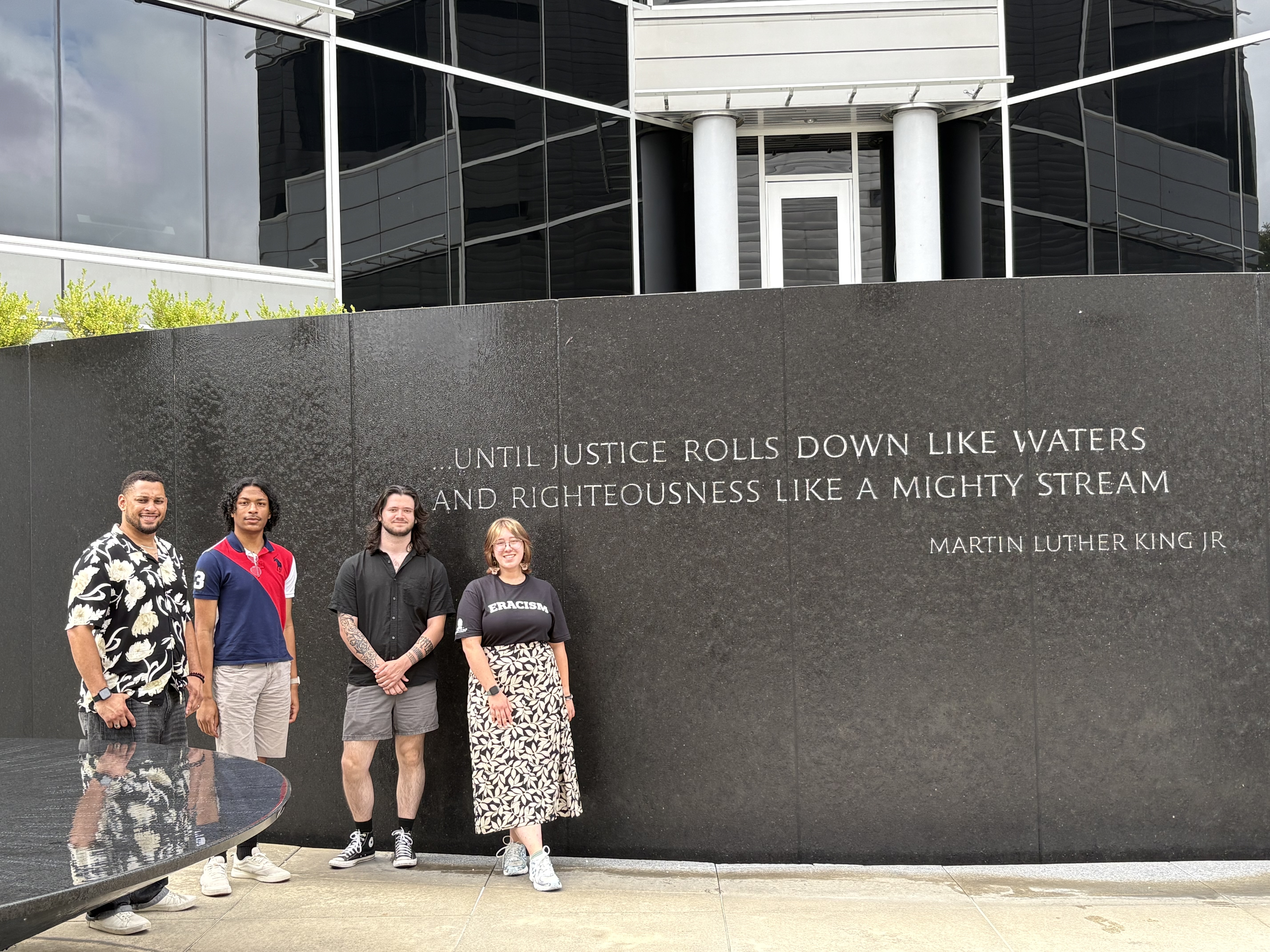 Four students stand in front of a memorial to Dr. King in front of a building in Montgomery. It is a water feature monument with the quote "Until justice rolls down like waters and righteousness like a mighty stream."