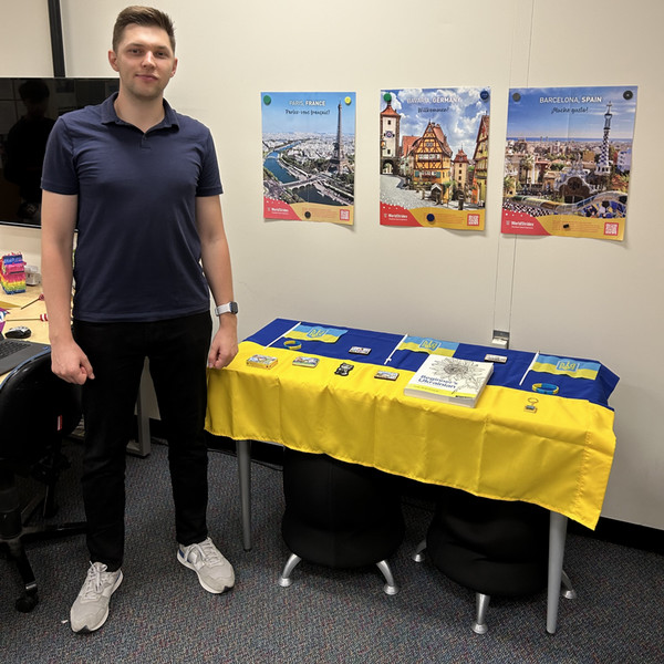 A young man stands in a room beside a table with the Ukrainian flag, photography of Ukraine on the wall, and informational pamphlets. 