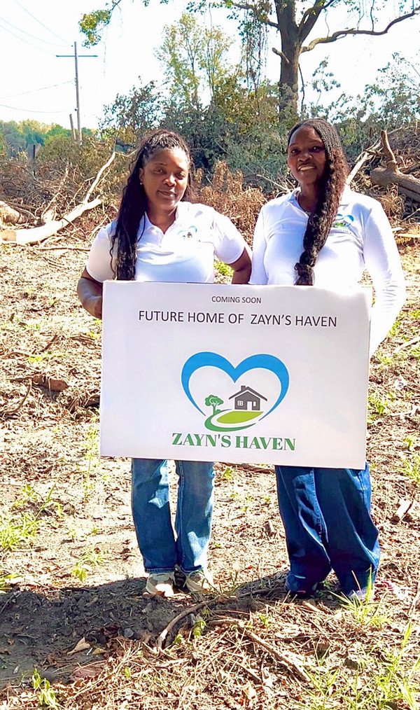 Two women standing behind a Future Home of Zayn's Haven yard sign
