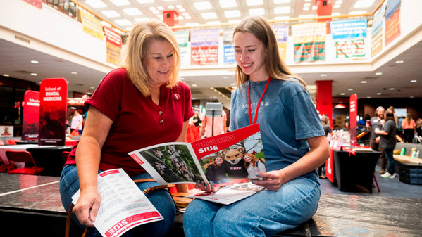 A mother and her high school aged daughter sit in the Goshen Lounge illuminated by skylight and are reading a booklet that reads Preview SIUE on the cover