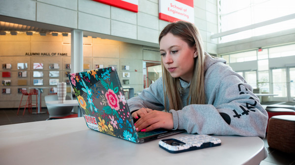 A student in the SIUE School of Engineering atrium leans into her studies, focused on a flower-covered laptop adorned with a Cougar sticker.
