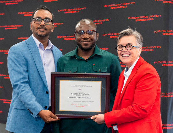 Two people flank thesis award winner as he holds his plaque and all face camera smiling