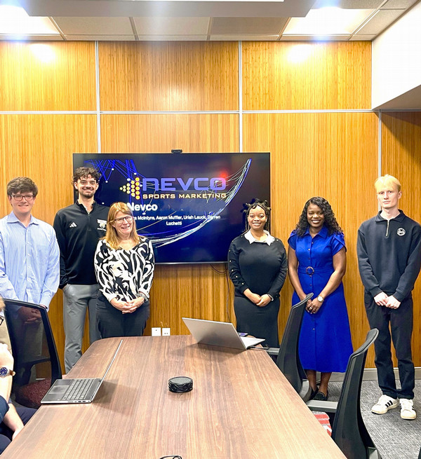 Group stands around conference table near TV monitor on wall