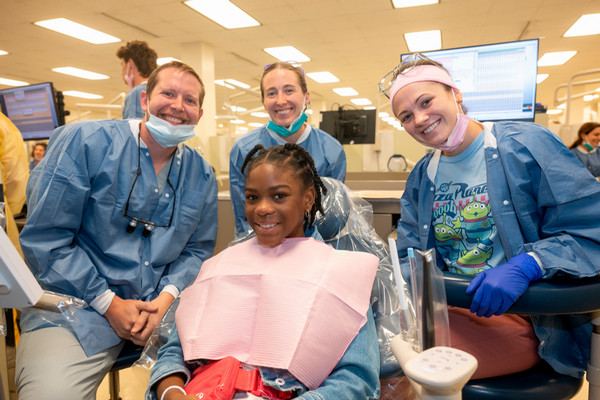 Gabrielle James beams from the dental chair, surrounded by three smiling members of the SIU SDM care team. Dressed in blue gowns, they share a joyful moment in a clinical room.