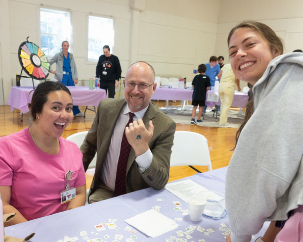 SIU SDM Dean Drukteinis smiles alongside two dental students at a table, proudly displaying a playful temporary tattoo of a smiley pirate on his hand amid colorful games and activities behind them.