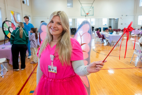 Zoey Lane bringing magic to Give Kids a Smile Day, smiling in bright pink scrubs, shimmering fairy wings and a matching tutu, with a red ribbon wand in hand as children enjoy activities behind her in a gymnasium.