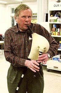 A man stands indoors playing a Bulgarian gaida (bagpipe), wearing a plaid shirt and dark trousers. He holds the bag under one arm and presses the chanter with both hands. Store shelves with products are visible in the background.