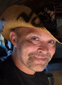 Candid photo of Brandon Triplett wearing a warm smile, kind eyes, and a cowboy hat with decorative spray paint lettering.