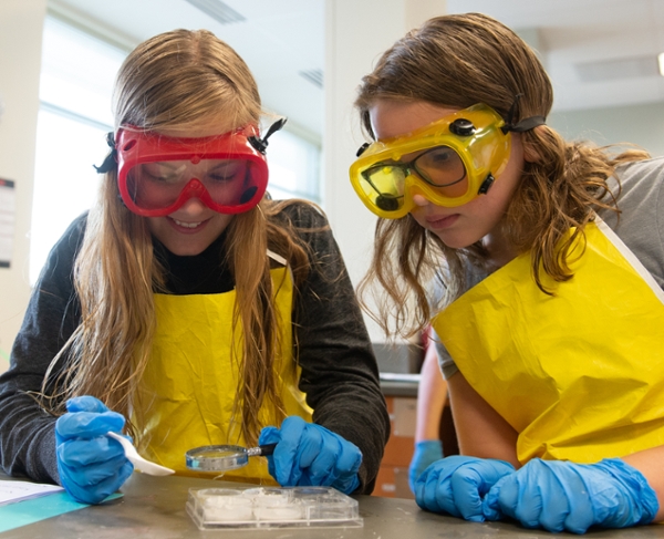 2 girls wearing eye protection looking at a petri dish with a magnifying glass