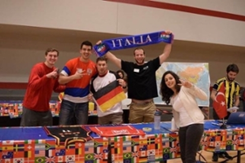 table with four male students behind and one female student in front, one male student is holding a sign that says Italia and another male student is holding the Italian flag