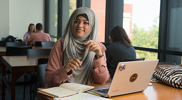 female Indian student smiling while sitting at study table with laptop in library